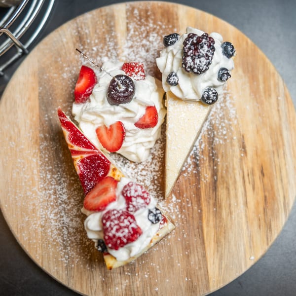 Three dessert slices with whipped cream and fresh berries, dusted with powdered sugar on a wooden board.