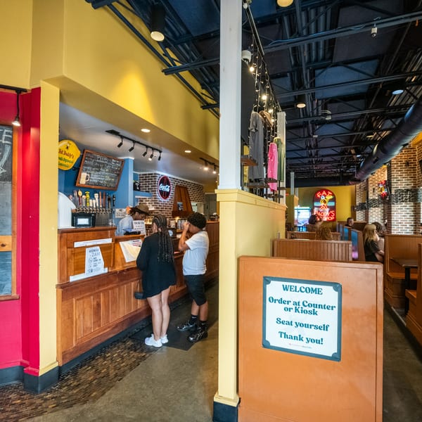 Customers order at a counter in a colorful restaurant with booths and exposed ceiling ducts.