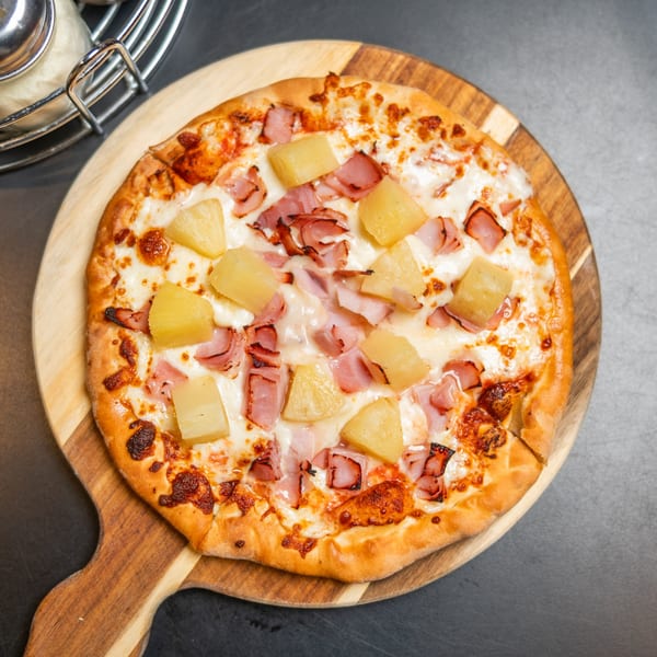 Hawaiian pizza with ham and pineapple on a wooden paddle, next to a cheese shaker.