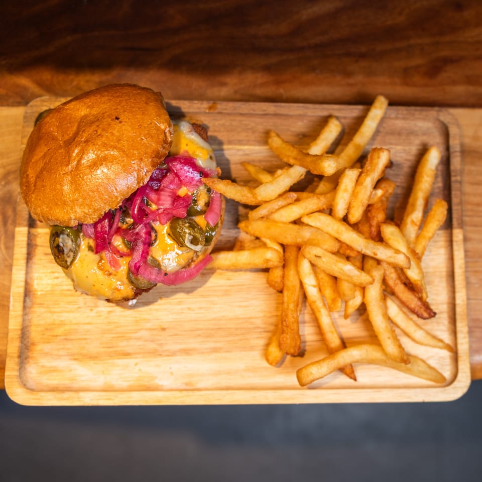 A cheeseburger with pickled onions and jalapeños, served with french fries on a wooden tray.
